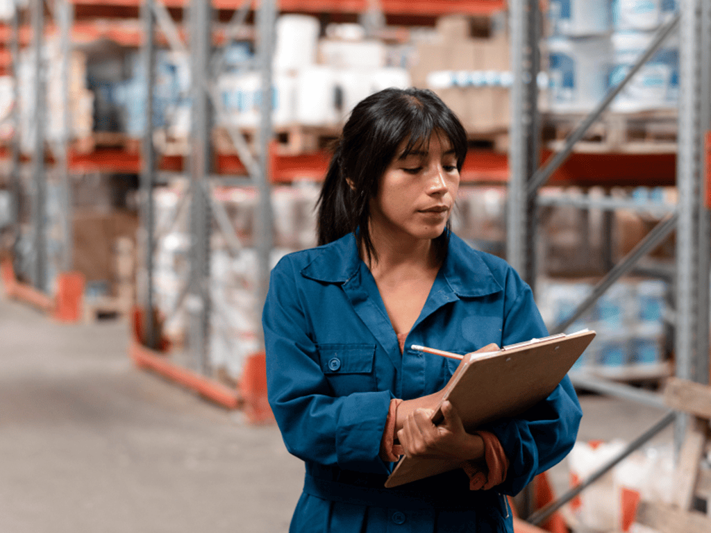 Trabajadora de logística con overol azul verifica inventario con portapapeles en bodega; inclusión laboral y aprendizaje con propósito.