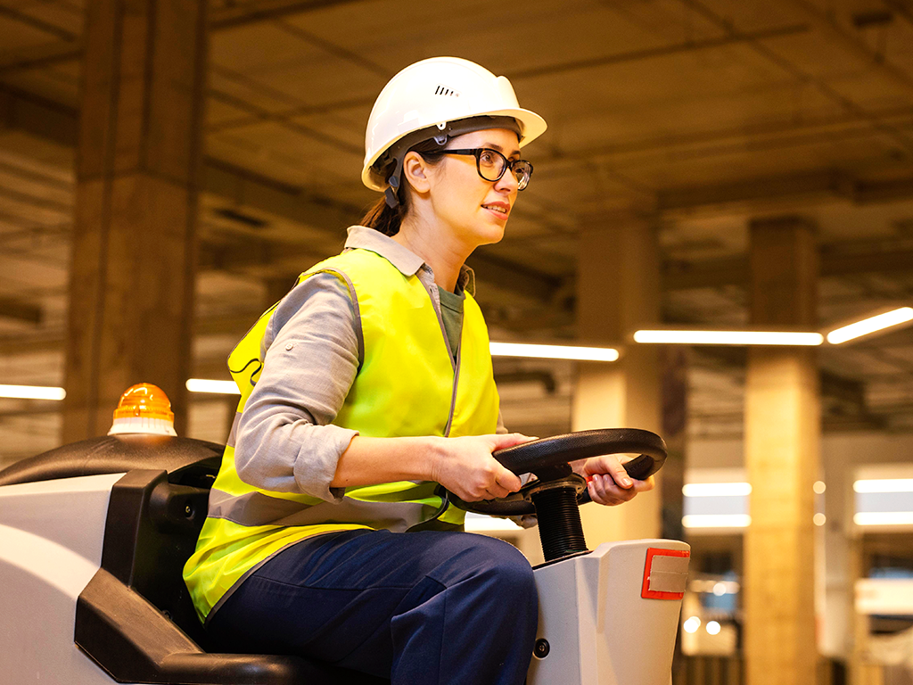 Operaria con casco y chaleco conduciendo maquinaria en bodega; formación práctica para mejorar seguridad y productividad.